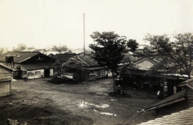 Black-and-white archive photograph of low buildings, a courtyard, trees and a tall chimney